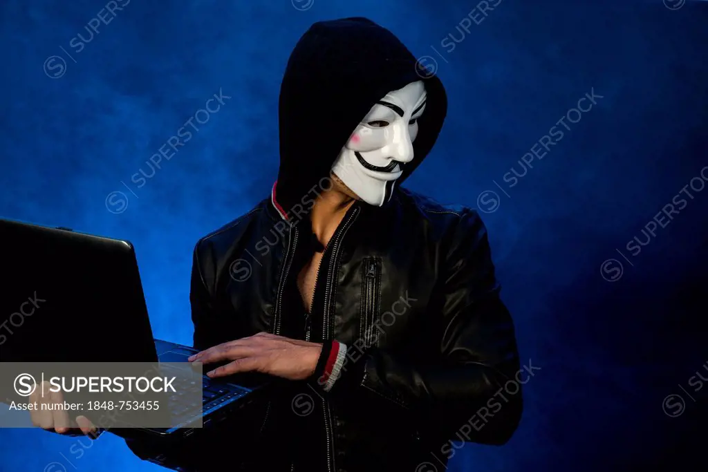 Young man wearing an anonymous mask with a hood and a black leather jacket, holding a laptop and looking to the side