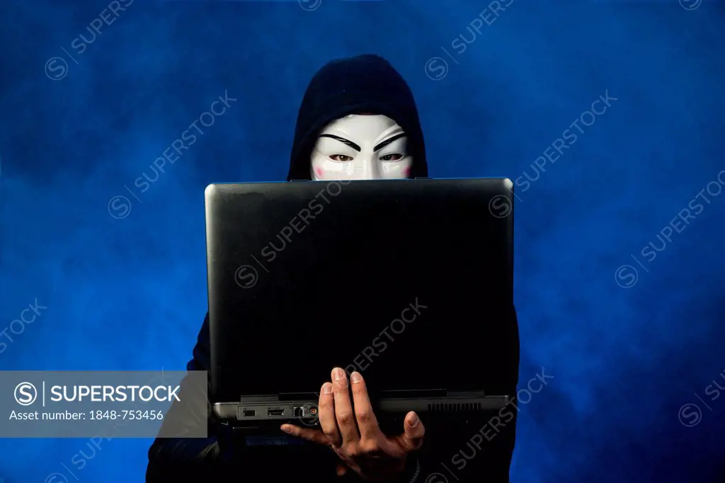 Young man wearing an anonymous mask with a hood, holding a laptop