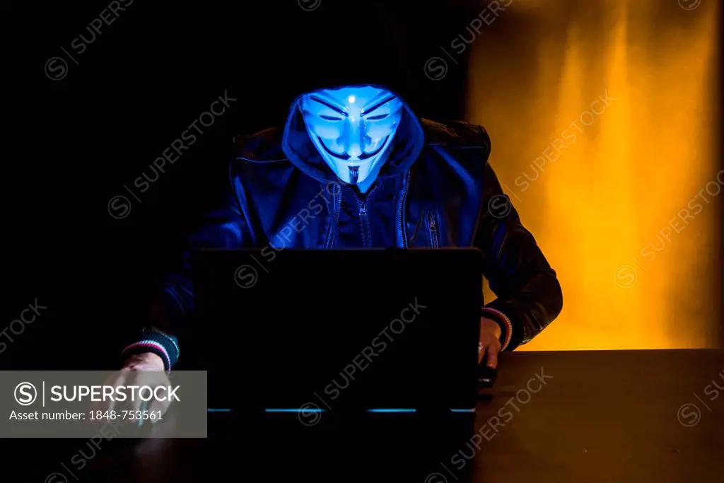 Young man wearing an anonymous mask and a hood sitting in front of a laptop