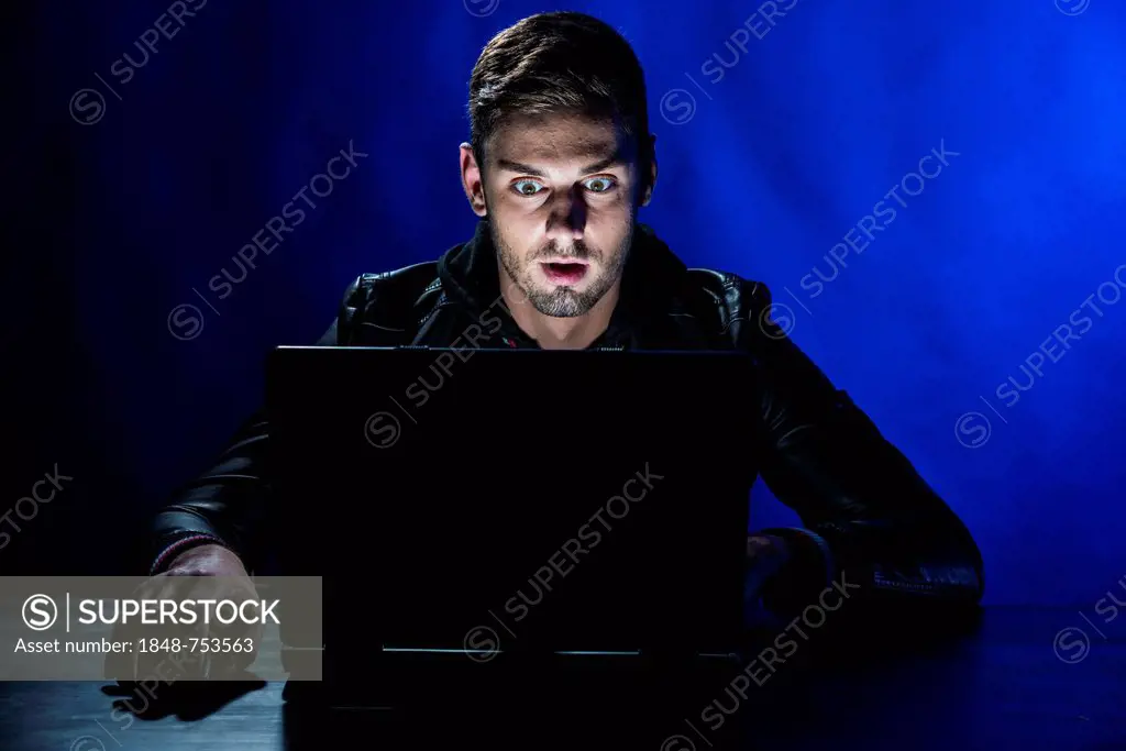 Young man sitting in the dark in front of a laptop, with a shocked facial expression