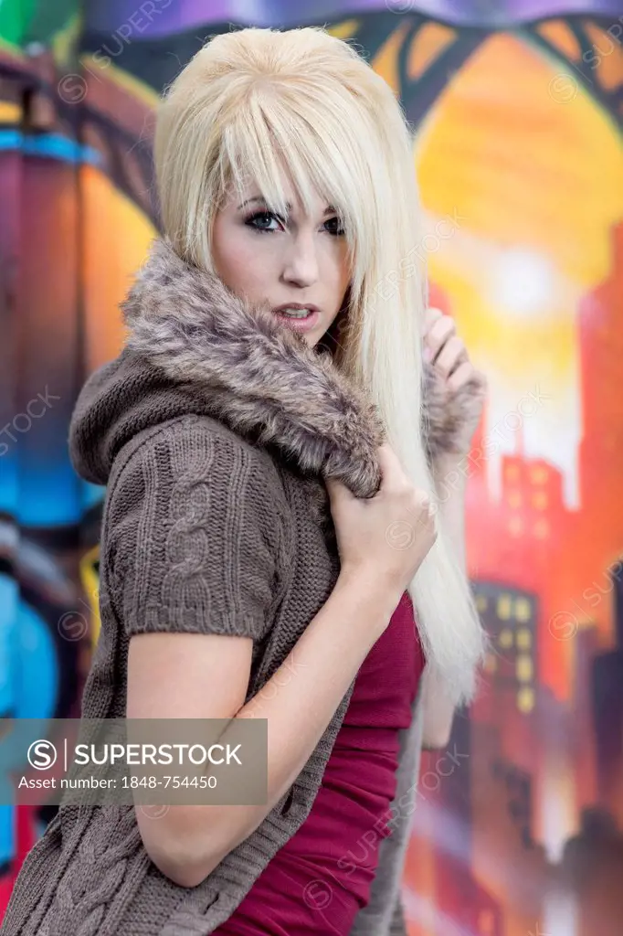 Young woman with long blond hair wearing a hooded jacket posing in front of a wall with graffiti, portrait