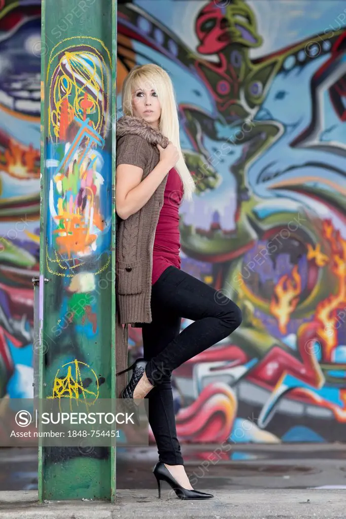 Young woman with long blond hair wearing a hooded jacket posing while leaning against a steel beam with graffiti in front of a wall with graffiti