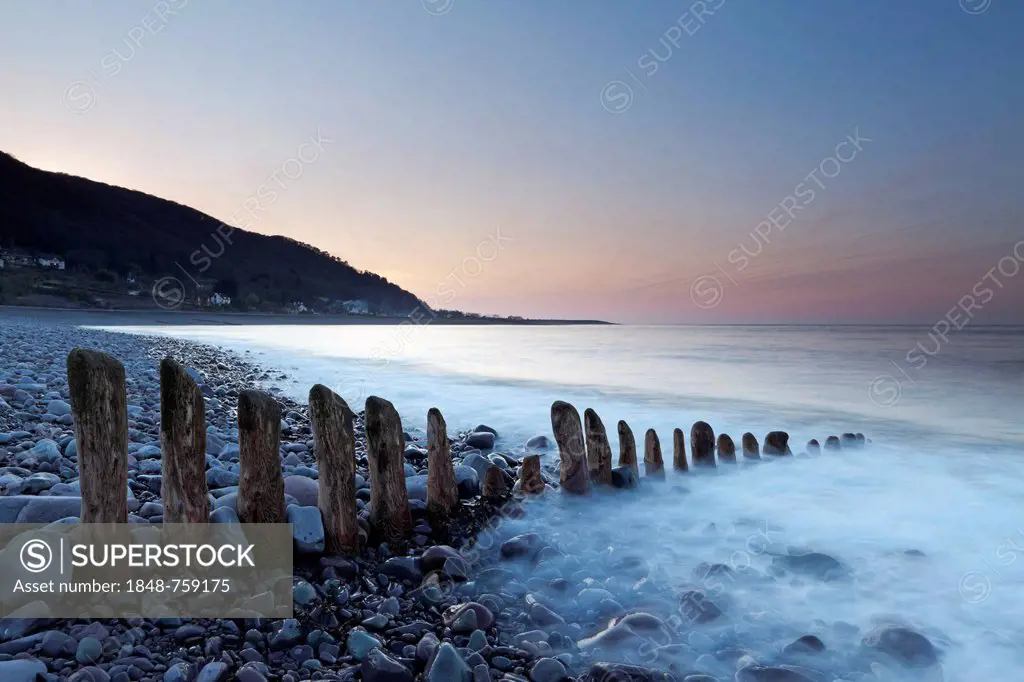 Waves washing over rocks, split by a groyne, at dusk, with the village of Porlock Weir at back, England, United Kingdom, Europe