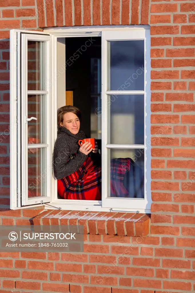 Young woman, 35 years, sitting with a cup of coffee and a blanket on the window sill of a lattice window in winter
