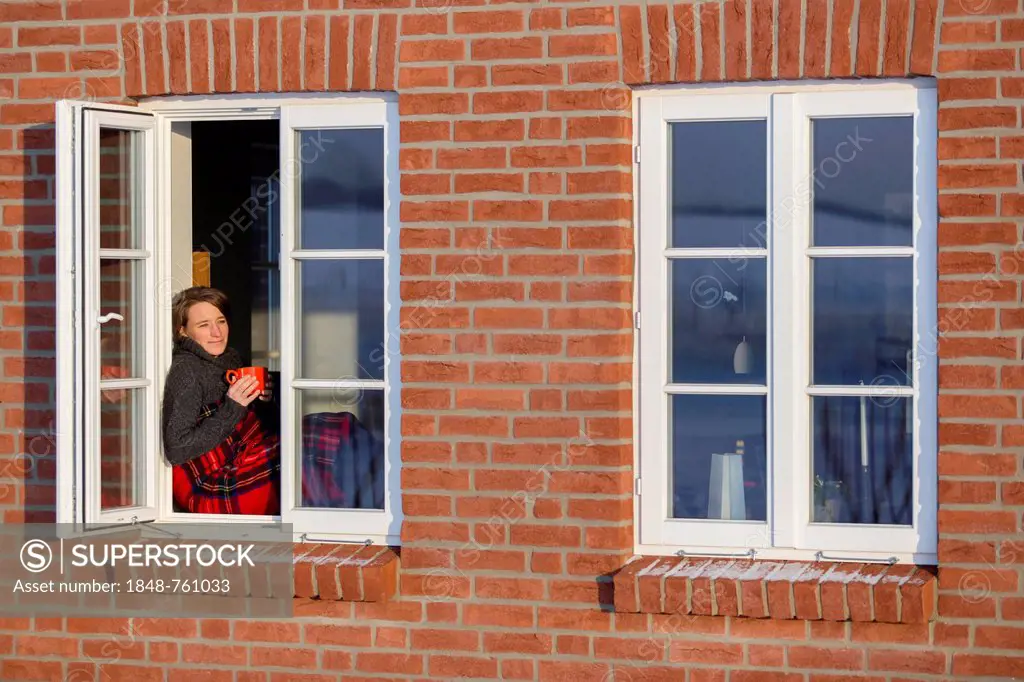 Young woman, 35 years, sitting with a cup of coffee and a blanket on the window sill of a lattice window in winter