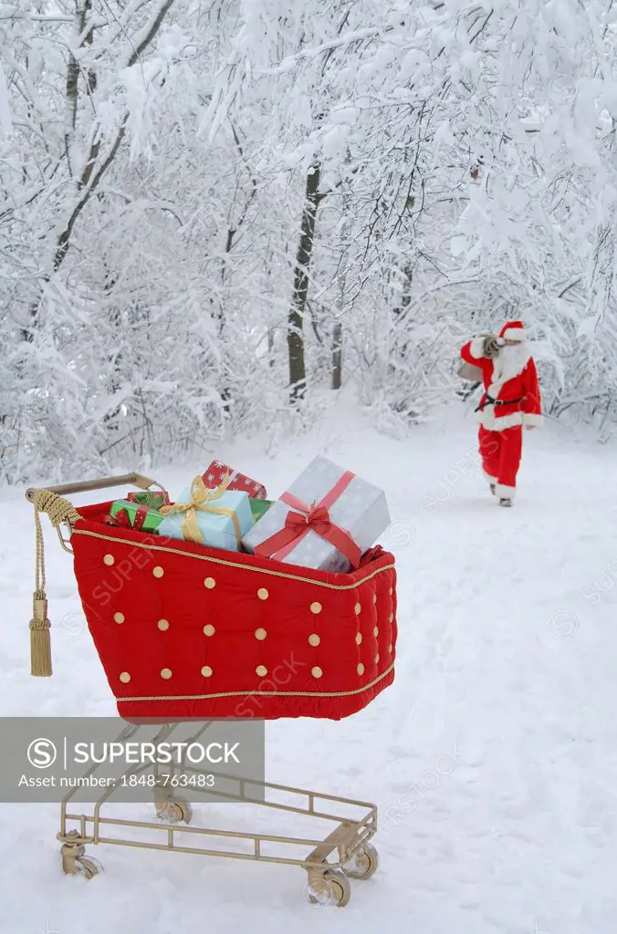 Santa Claus in a snowy forest with a shopping cart with red velvet and gifts in the foreground