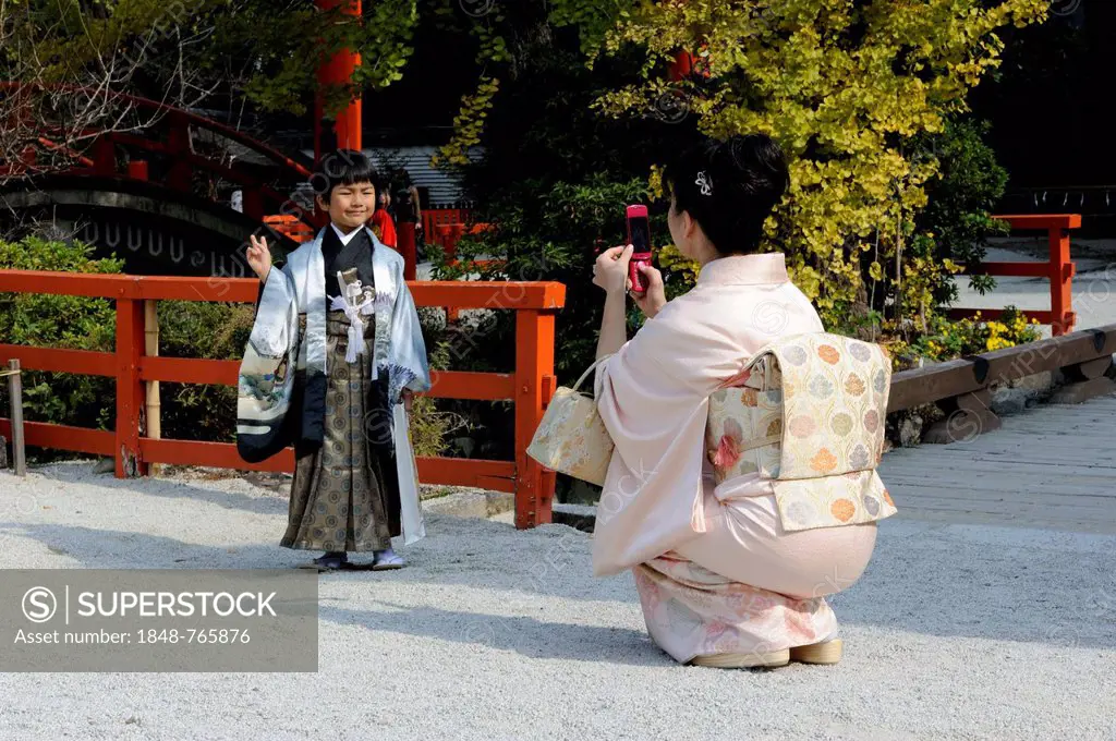 Japanese boy wearing an ornate Kimono, Hakama culottes and a Haori, a kimono-like jacket, with a victory hand gesture, his mother is wearing a kimono ...