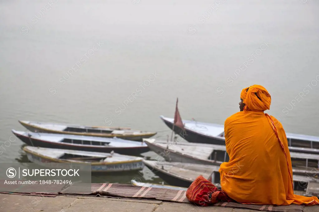 Brahmin or Hindu priest sitting on a blanket on the banks of the Ganges River with several boats tied to the shore