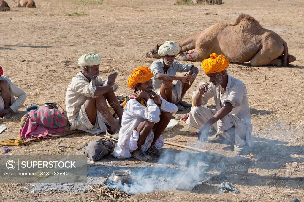 Four elderly Indian men with turbans and wearing the traditional Dhoti garment squatting on the ground, food is being prepared over an open fire, Push...