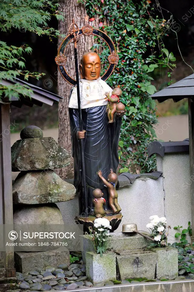 Buddhist Jizo statue with bib and halo, the patron god of the children, with a child in his arms and children at his feet, next to a stone lamp