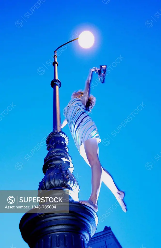 Woman wearing a summer dress standing on street lamp and holding hershoes in the air, blue hour