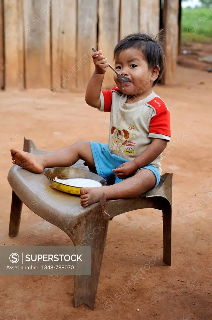 Boy, 3, sitting on a broken chair and eating porridge, in a community of the Guarani Indians