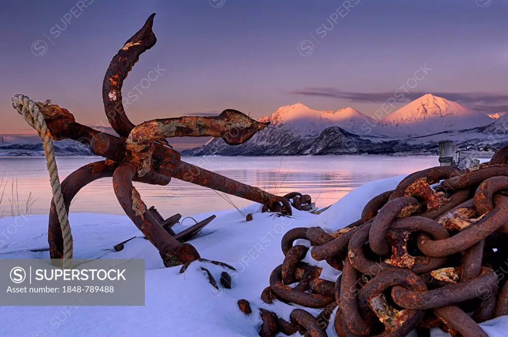 Rusty anchor in front of a fjord with a mountain range in the evening light