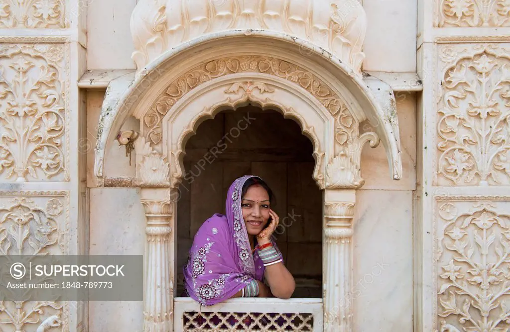 Young friendly Indian woman wearing a headscarf looking out of a window opening of an ornate marble facade, Rat Temple of the goddess Karni Mata, Karn...