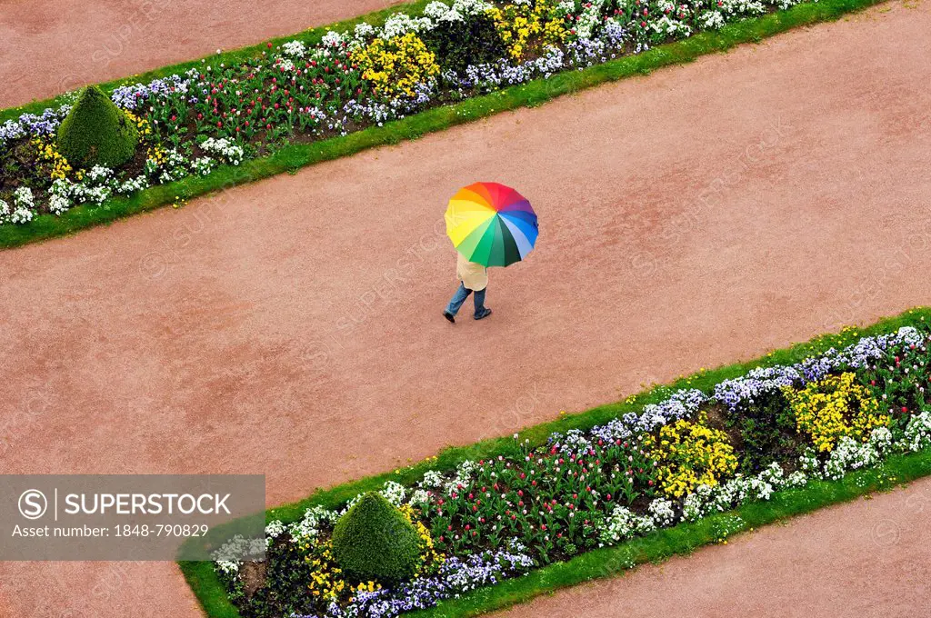 Person with an umbrella in rainbow colours on a red dirt road between flowerbeds in the Palace Gardens, Stadtschloss City Palace