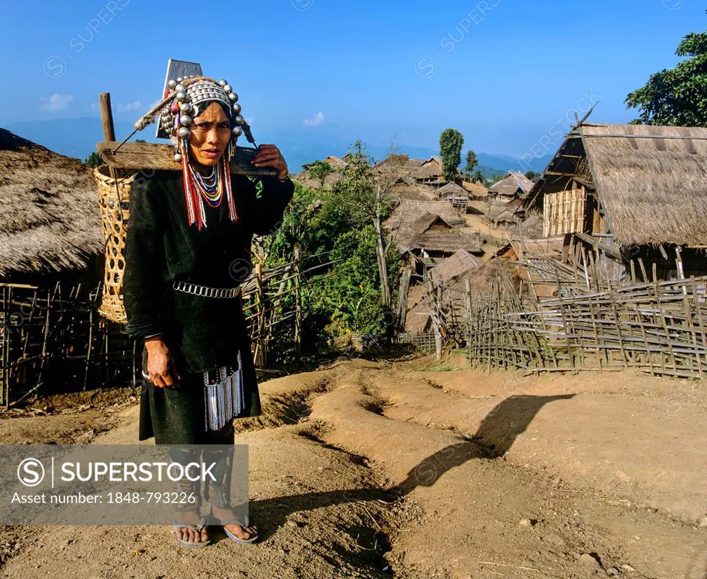 Akha woman in traditional costume with a pannier, in a mountain village with bamboo huts