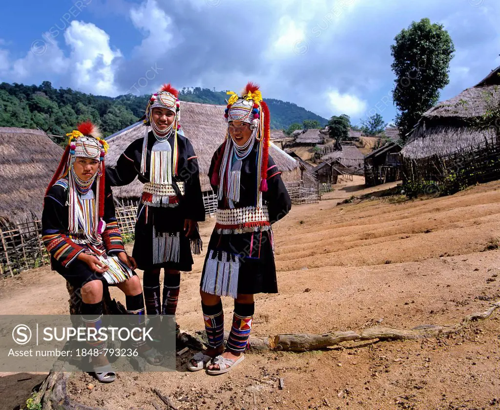 Three Akha girls in a mountain village, in traditional costume and headdress, bamboo huts with thatched roofs at back