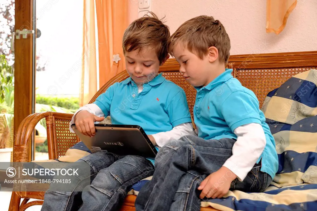 Two boys, twins, sitting side by side on a bench in a living room playing on a tablet computer