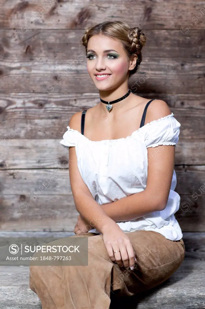 Young woman wearing a white dirndl top and traditional leather pants sitting on a wooden bench, dirndl look