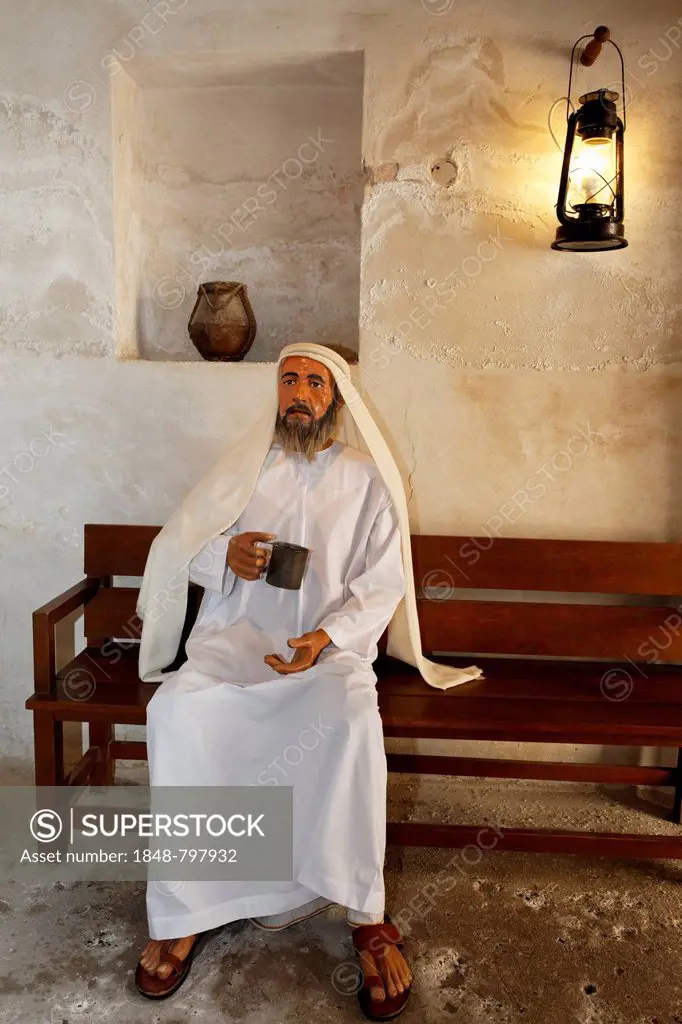 Arab wearing a traditional white dishdasha robe sitting on a bench with a cup of water, life-size figure, Al-Ahmadiya School Museum, Dubai, United Ara...