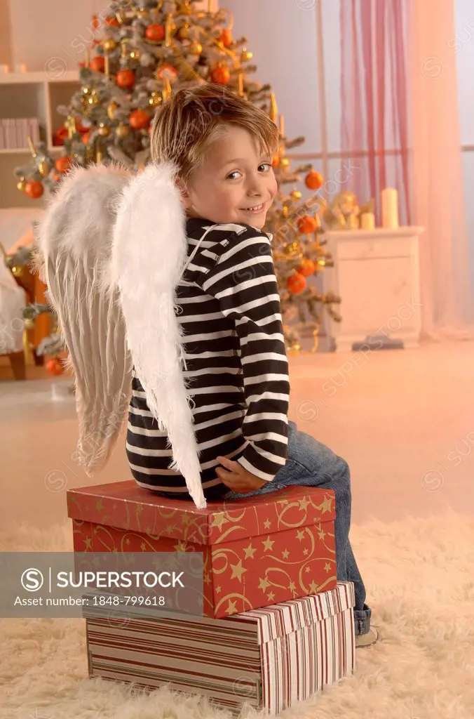 Boy wearing angel wings sitting on gifts in front of a Christmas tree