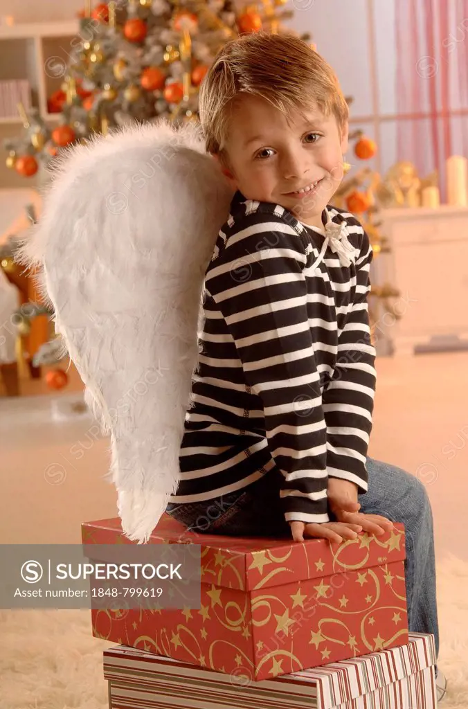 Boy wearing angel wings sitting on gifts in front of a Christmas tree