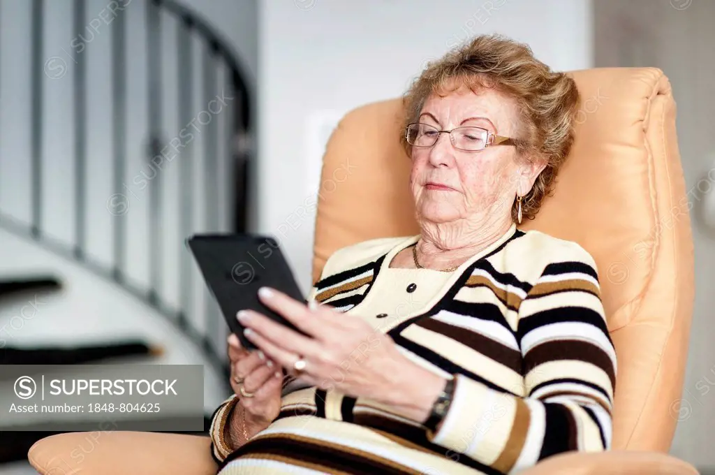 Elderly woman sitting in an armchair and reading an eBook, Germany, Europe