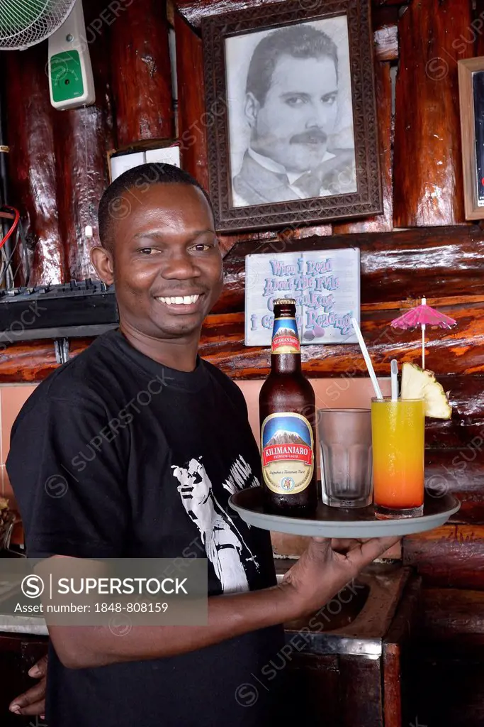 Waiter with Kilimanjaro beer and a cocktail in front of a picture of Freddy Mercury, the late singer of the rock band Queen who was born in Zanzibar, ...