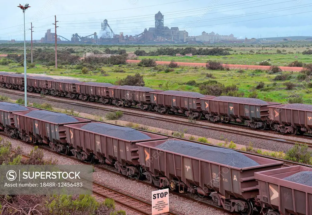 Freight train loaded with iron ore, at the entrance to the ore-loading port, at the back the Saldanha Steel steelworks, Saldanha, Western Cape, South ...