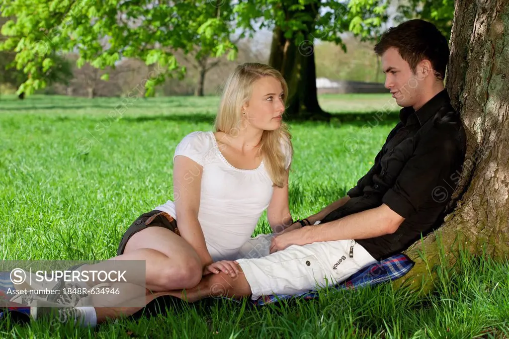 Young couple sitting on a picnic blanket and leaning against a tree in a park in spring