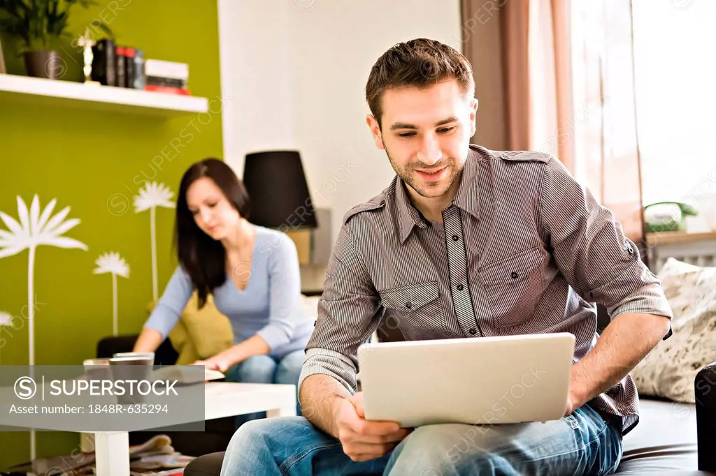Young man at home, with a laptop, young woman at back