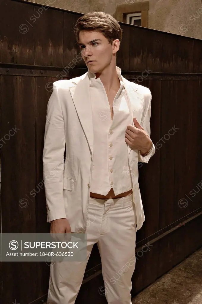 Fashion photo of a young man wearing a white suit standing in front of a wooden fence