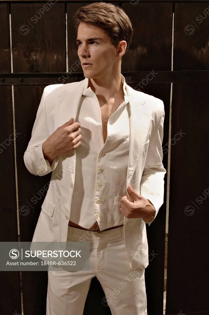Fashion photo of a young man wearing a white suit standing in front of a wooden fence