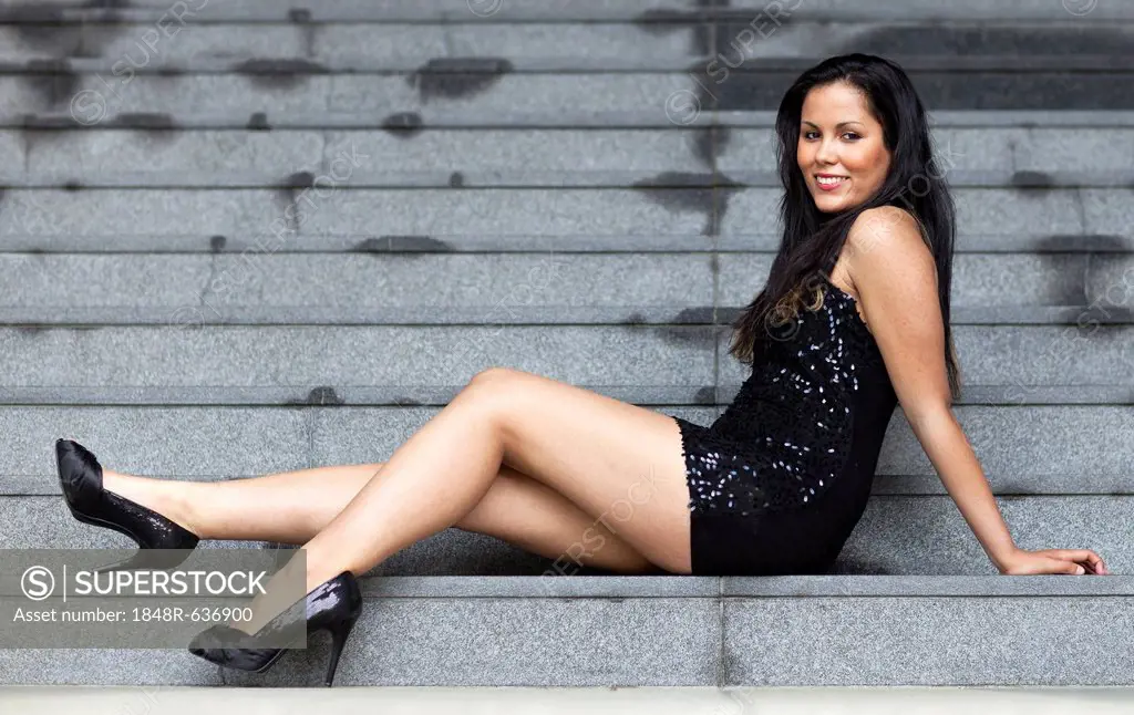 Young woman with long dark hair, short dress and high heels posing sitting on stone steps