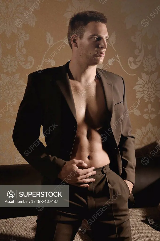 Bare-chested young man wearing a suit standing in front of fabric wallpaper in a living room