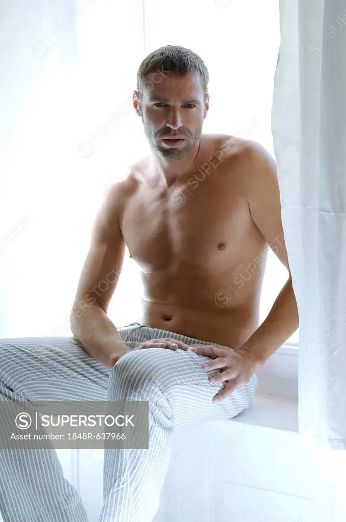 Bare-chested man sitting in bedroom suffused with light