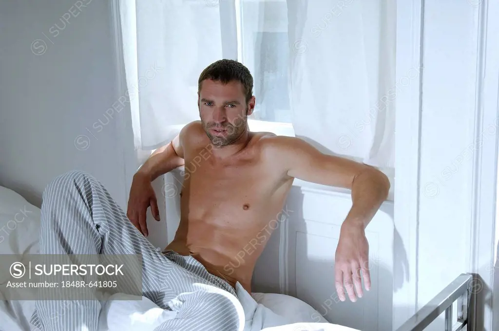 Bare-chested man sitting on bed in bedroom suffused with light