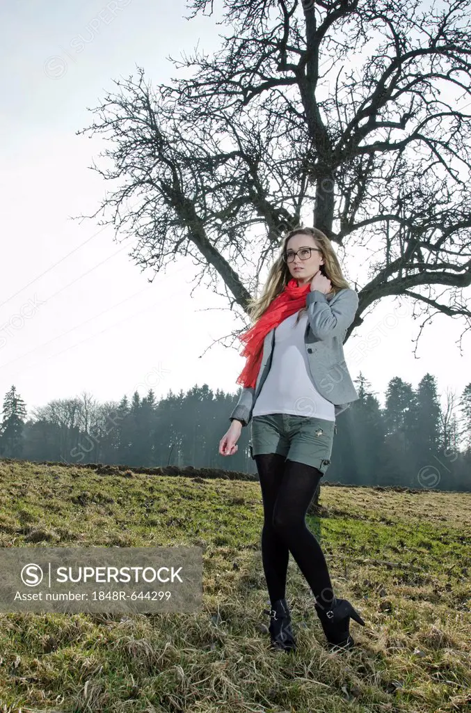 Young woman, 25, wearing a red scarf on a meadow in front of a bare tree