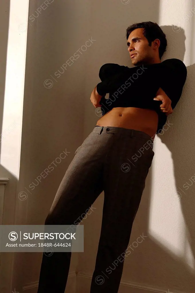 Young man leaning against apartment wall, taking off his sweater