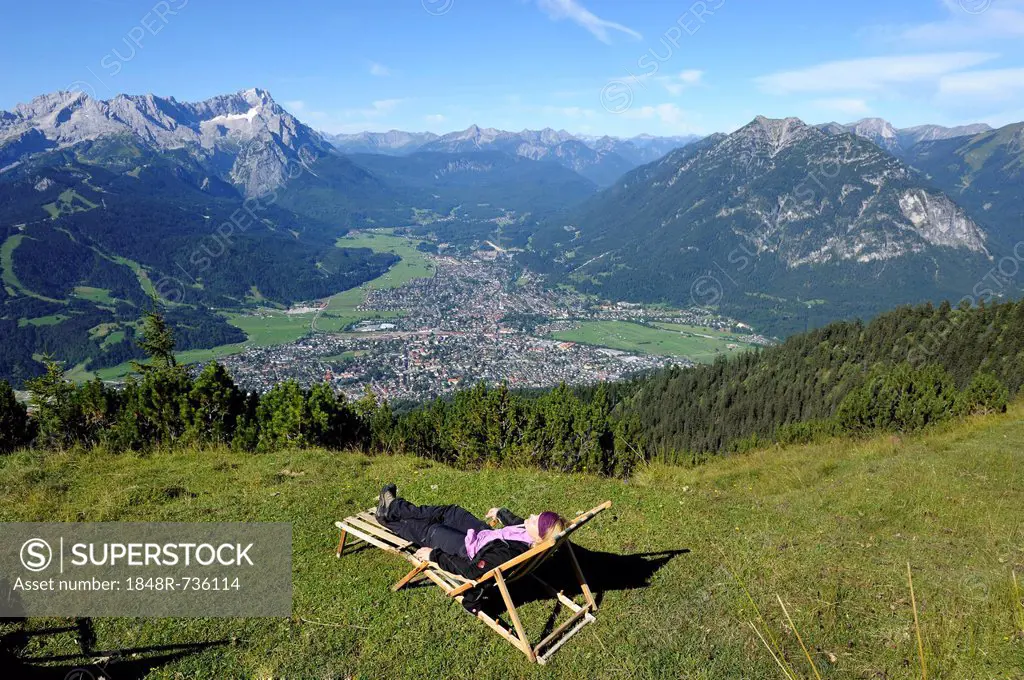 Young woman in a deck chair, view of Garmisch-Partenkirchen, Zugspitze mountain, Jubilaeumsgrat ridge and Alpspitze mountain as seen from Wank mountai...