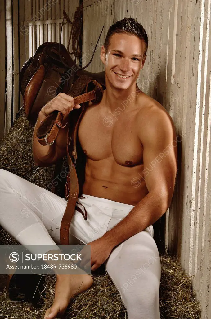Man with a naked torso wearing long underwear in a horse barn sitting on bales of hay