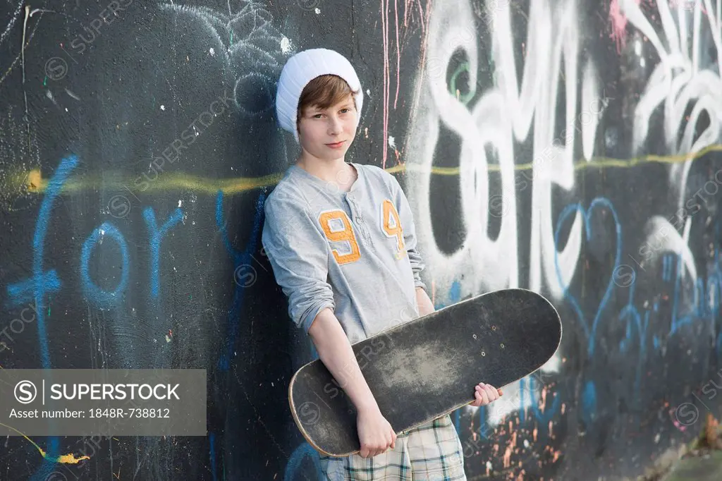 Boy holding a skateboard standing in front of a wall with graffiti