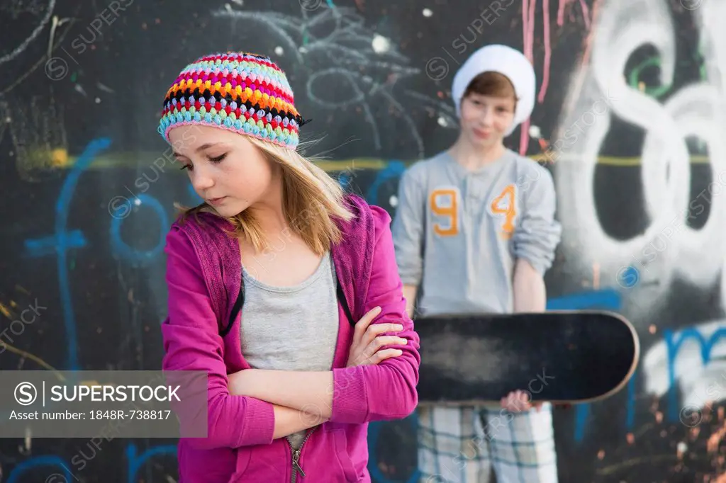 Girl and a boy holding a skateboard standing in front of a wall with graffiti