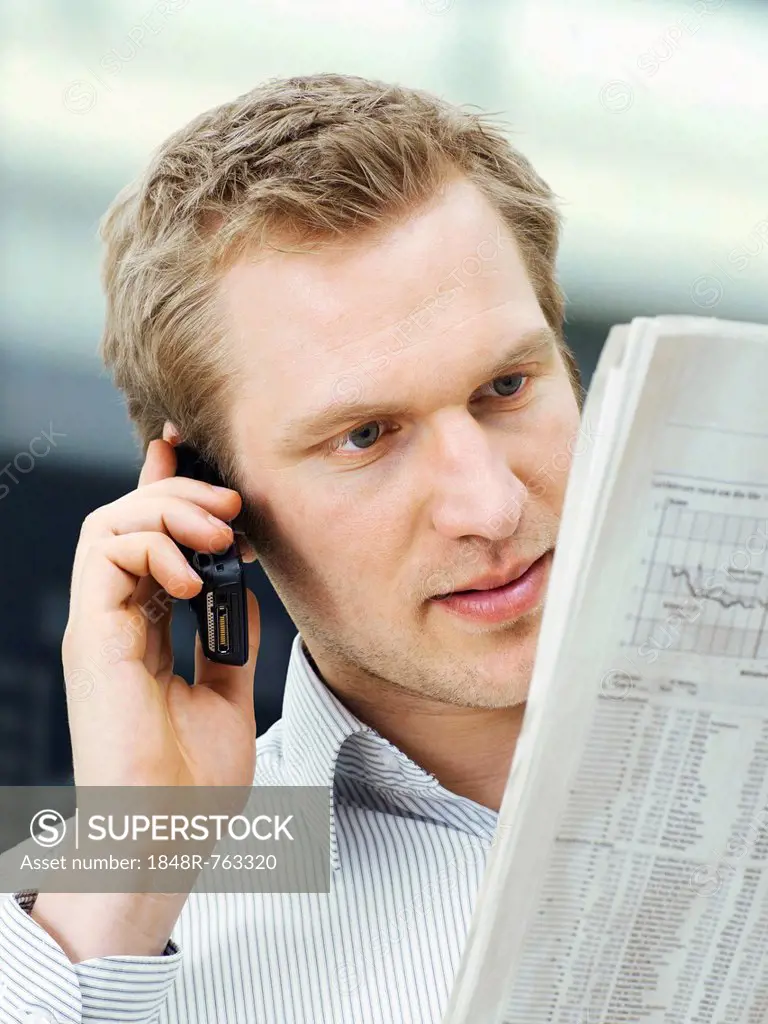 Businessman reading a newspaper while using a mobile phone, concentrating