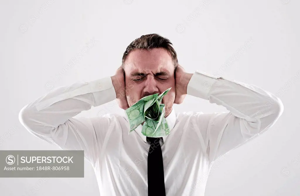 Young, desperate man wearing a shirt and a tie holding his hands over his ears, has 100-euro notes stuffed into his mouth