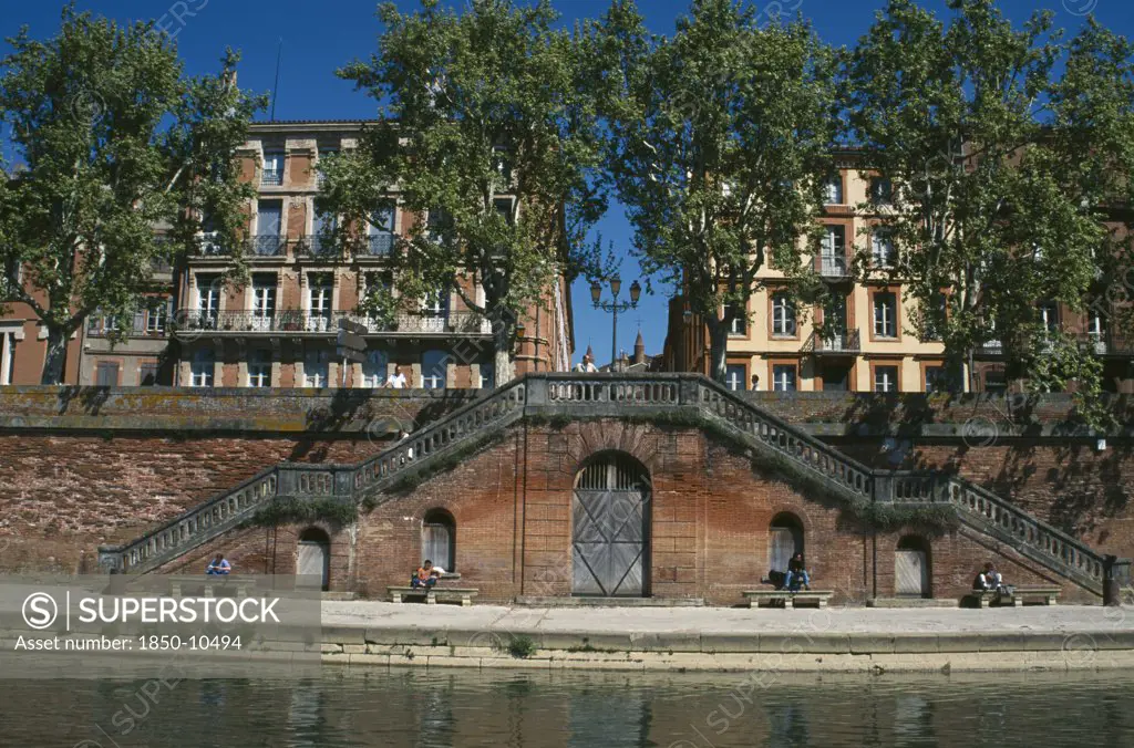 France, Midi Pyrenees, Haute Garonne, Toulouse.  Red Brick Steps Leading Down To The River Garonne From Street Level With Facades Of Buildings Partly Seen Through Trees Behind.  People Sitting In The Sun.