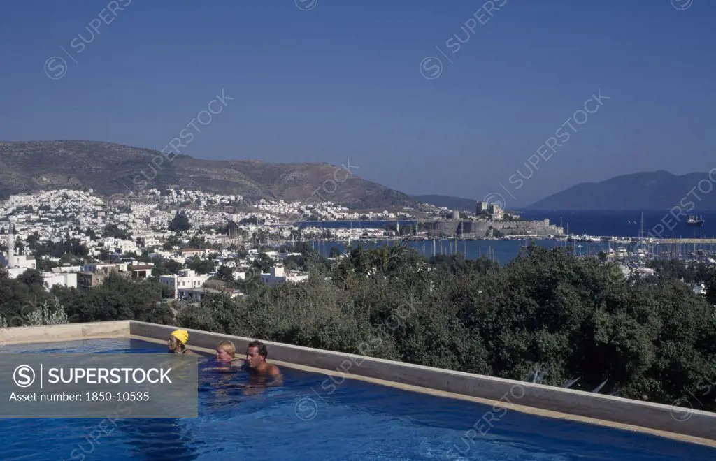 Turkey, Aegean Region, Mugla, Bodrum.  People In Swimming Pool Of Antik Theatre Hotel In Foreground With View Over Resort To Fifteenth Century Castle Of St Peter Beyond.