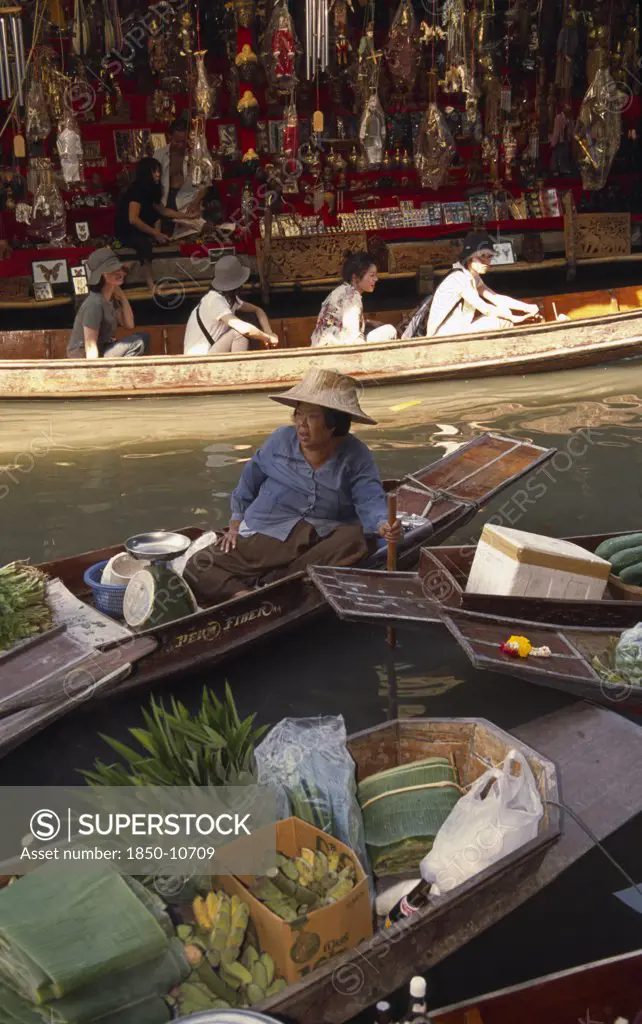 Thailand, South, Bangkok, Damnoen Saduak Floating Market With An Old Female Fruit Vendor In Her Canoe And Tourists In A Canoe Behind In Front Of A Tourist Goods Stall