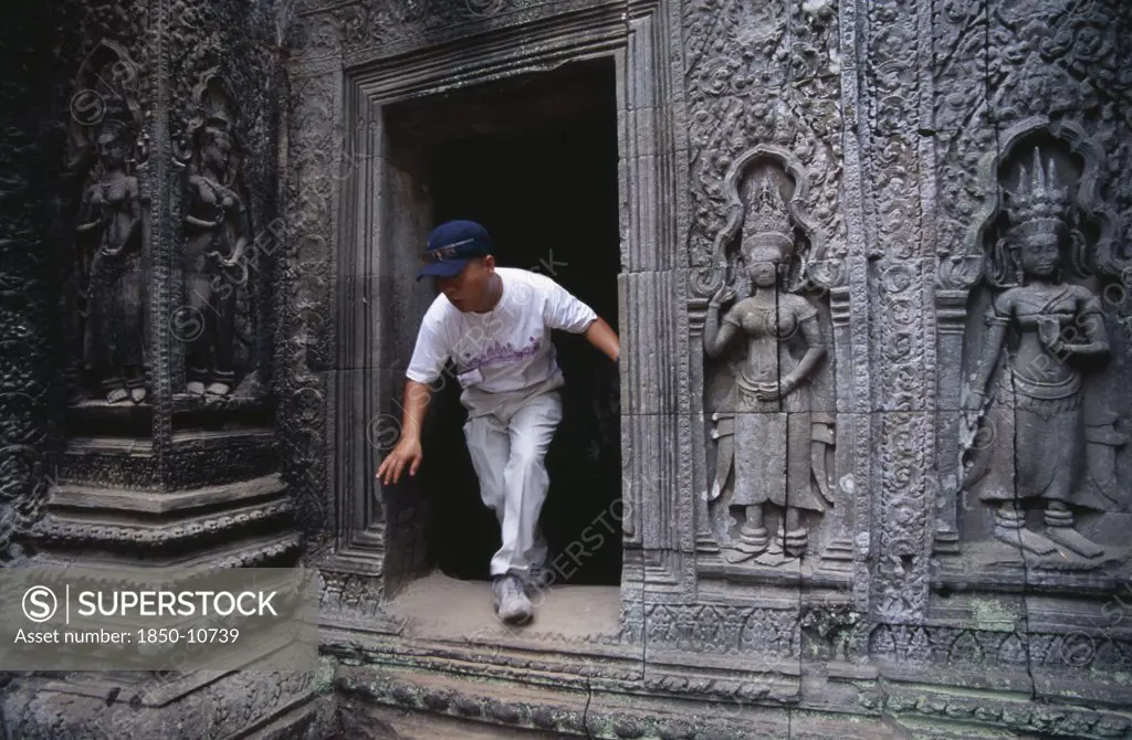 Cambodia, Siem Reap Province, Angkor, Ta Prohm.  Visitor Climbing Through Doorway Of Twelth Century Buddhist Temple With Exterior Wall Decorated With Bas Relief Carvings.