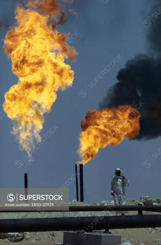 Saudi Arabia, Industry, Flaming Gas On Oil Field With Man Standing Behind Pipe In Foreground.